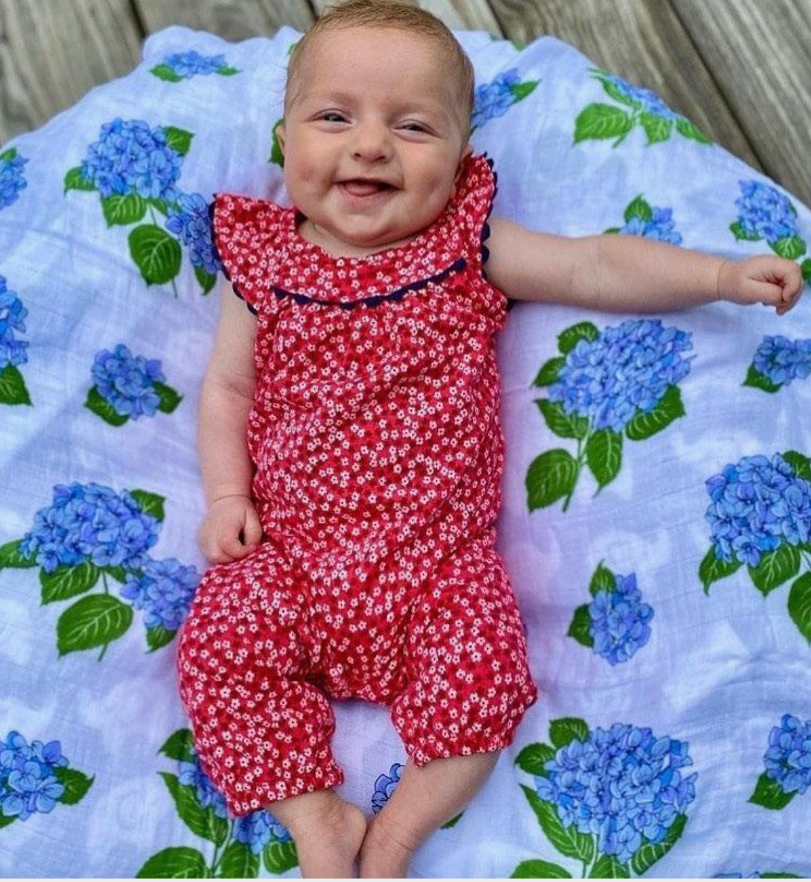 Baby in a red floral outfit lying on a Little Hometown Brand  blue hydrangea patterned swaddle blanket