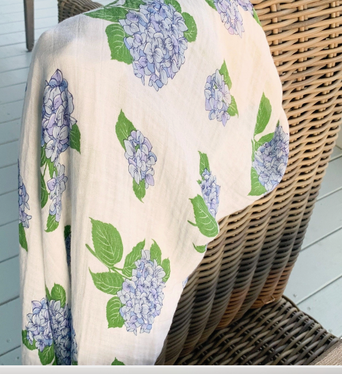 Little Hometown hydrangea swaddle blanket showing full floral print draped over a wicker chair on a wooden floor.