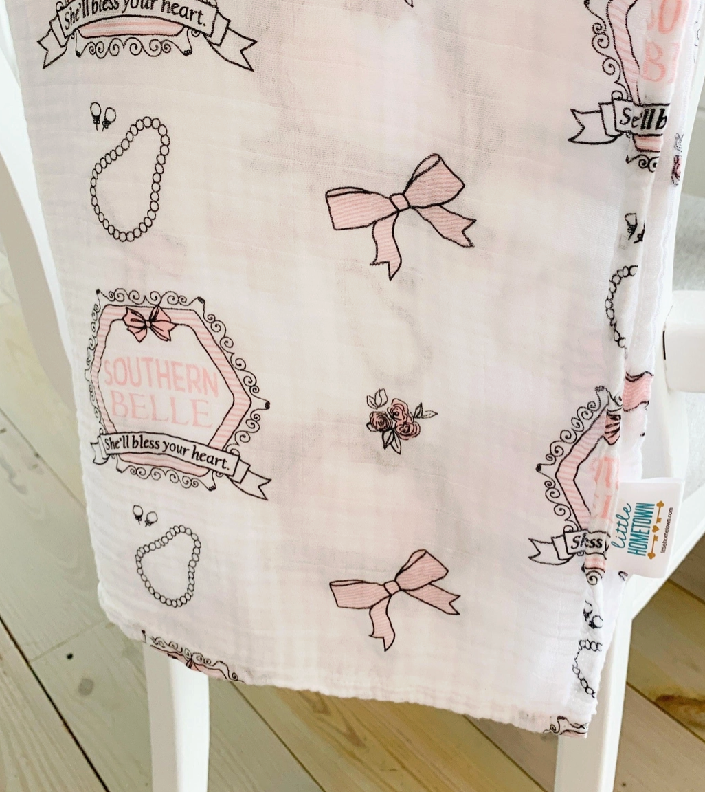 Little Hometown Brand Southern Belle Baby swaddle blanket with decorative pink and gray patterns and text draped over back of chair with wood floor. 