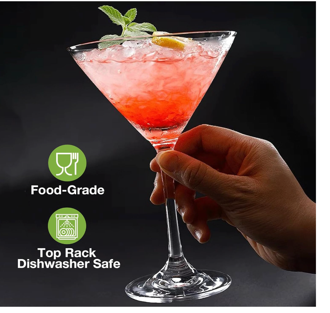 A hand holds a stemmed Huang brand Acrylic martini glass filled with a pink cocktail and ice, with a mint leaf and lemon garnish. Green icons on the left read 'Food-Grade' and 'Top Rack Dishwasher Safe' on a dark background.