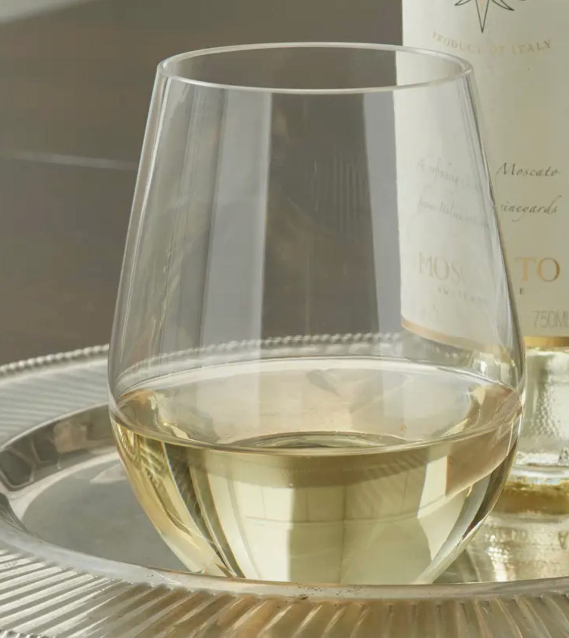 A clear stemless huang Acrylic wine glass containing white wine, set on a silver tray with a wine bottle partially visible in the background.