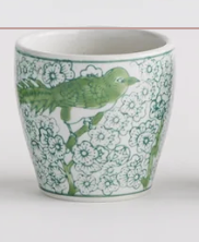 A white Napa Home and Garden Brand ceramic cup decorated with a green bird perched on a flowering branch, shown against a plain white background.