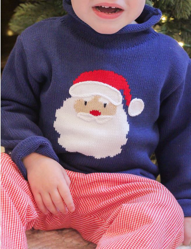 A close-up of a child wearing a navy blue cotton knit sweater with a large knitted Santa face on the front, paired with red-and-white gingham pants.