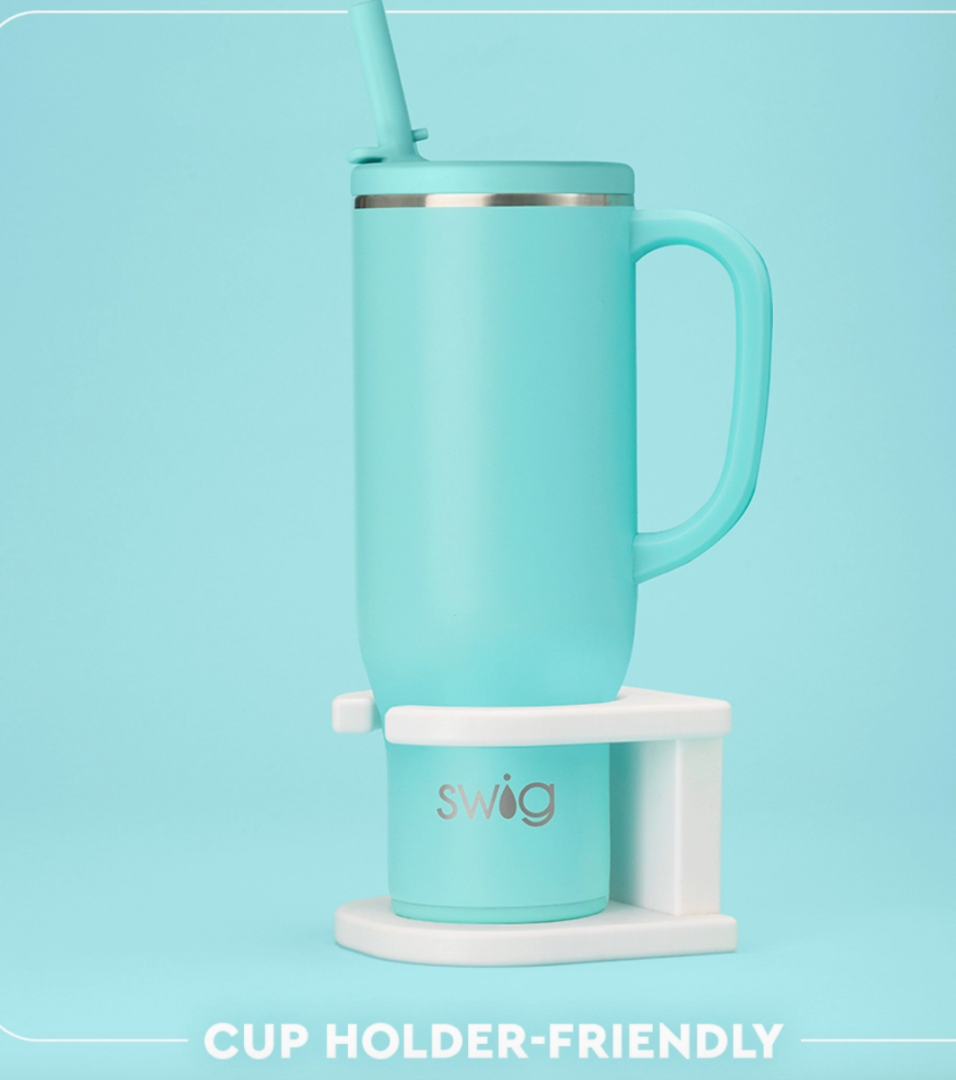 A light aqua insulated travel tumbler with a matching lid and straw, shown on a white cup holder stand against a light aqua background. The tumbler has a silver band near the rim and the word 'swig' printed on the holder.