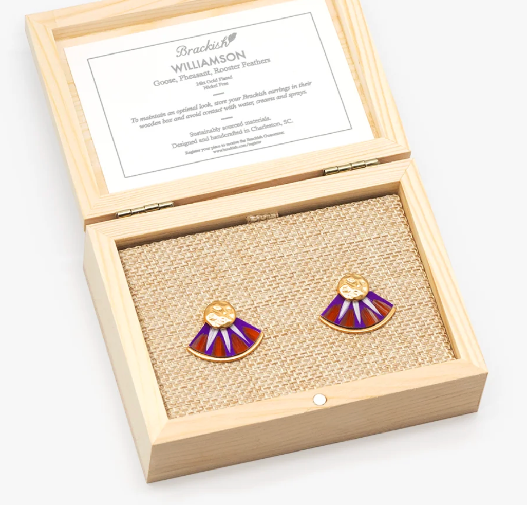 A pair of gold, orange and purple fan-shaped feather earrings displayed inside an open light wood presentation box with a branded insert card.