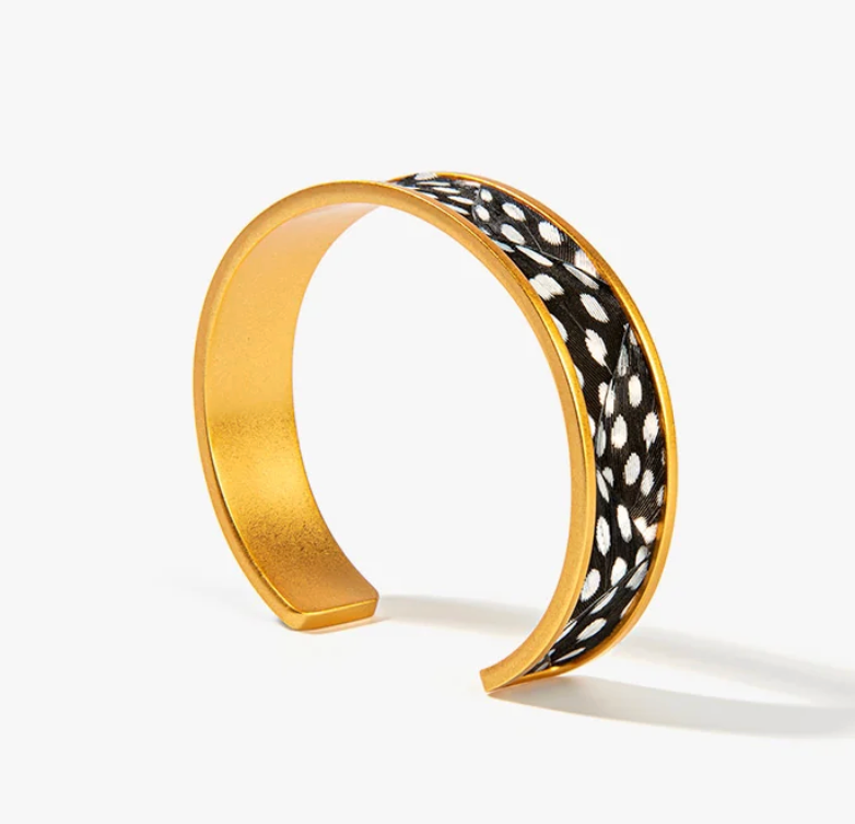 A gold-toned open cuff bracelet featuring black and white feathers set in gold outer band with a photographed on a clean white background.