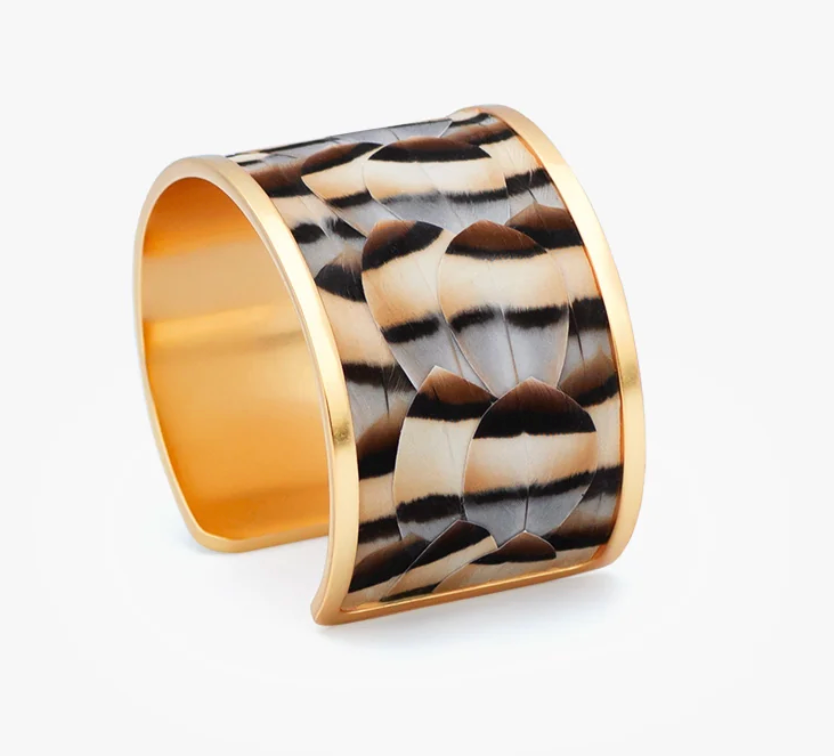A wide open cuff bracelet with a polished gold-tone metal border and an inlay of alternating brown, black, and cream feathers, shown on a light gray background.