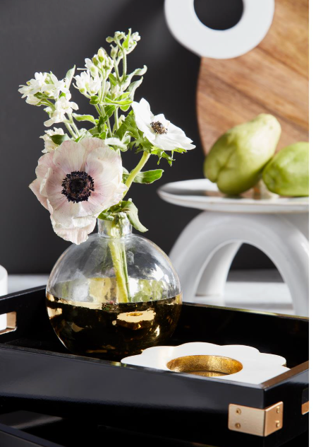 A clear glass vase with a reflective gold base holding white and pale pink flowers, styled on a black tray with a white flower-shaped dish underneath. A white pedestal holds green pears in the background against a dark wall.
