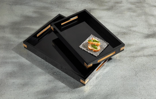 Two black rectangular serving trays with gold-toned handles and corner accents, arranged on a gray textured surface. One tray holds a small decorative dish with a piece of toast and greens.