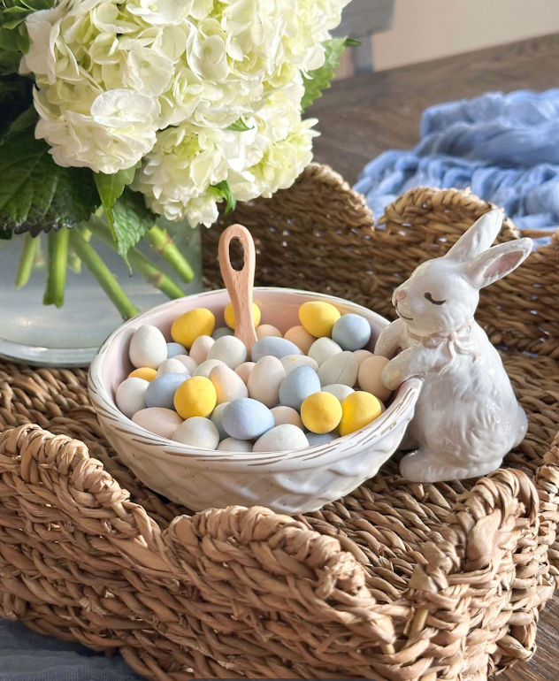 A decorative ceramic bowl filled with pastel Easter eggs sits on a woven wicker tray next to a white bunny figurine. The scene includes white flowers in the background and a soft blue textile, creating a styled home decor setting.
