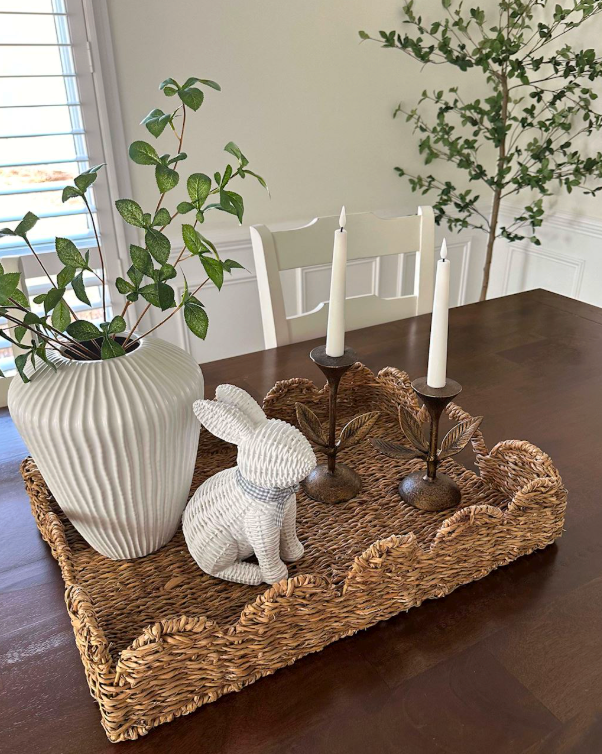 A woven rectangular table tray with a scalloped edge sits on a dark wood dining table. The tray is styled with a large white ribbed ceramic vase holding green leafy stems, a white knitted bunny figurine, and two bronze-toned candlesticks with white taper candles.