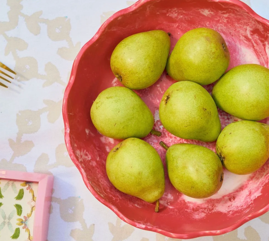Seven green pears arranged in a pink Laura Park brand scalloped bowl on a patterned white tablecloth, with a partial pink frame and a gold fork visible at the edges.