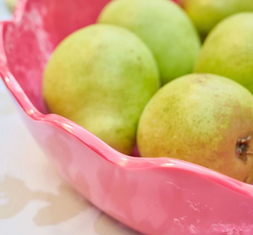Green pears in a Laura Park Designs brand scalloped pink resin bowl on a light patterned surface.
