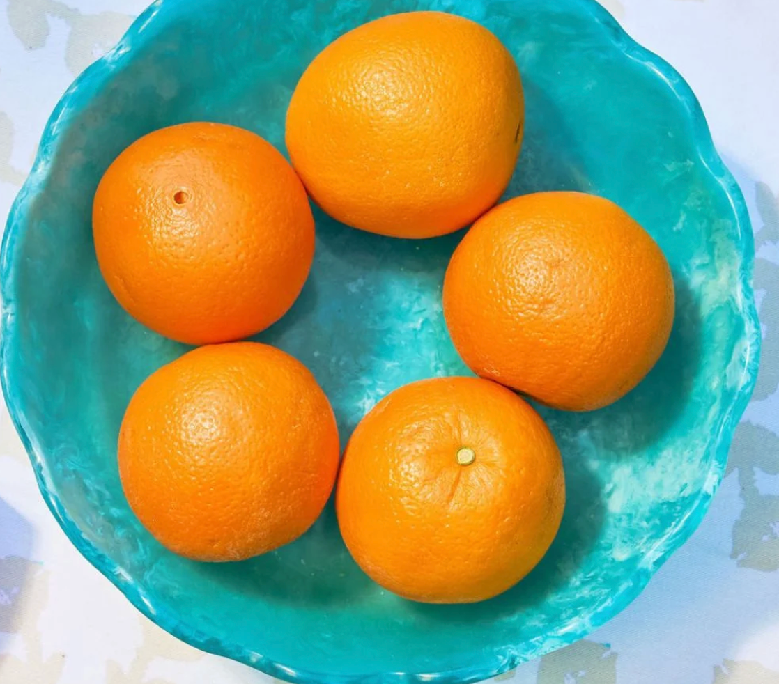 Five whole oranges arranged in a turquoise marbled resin bowl with scalloped edge on white background. Laura Park Brand