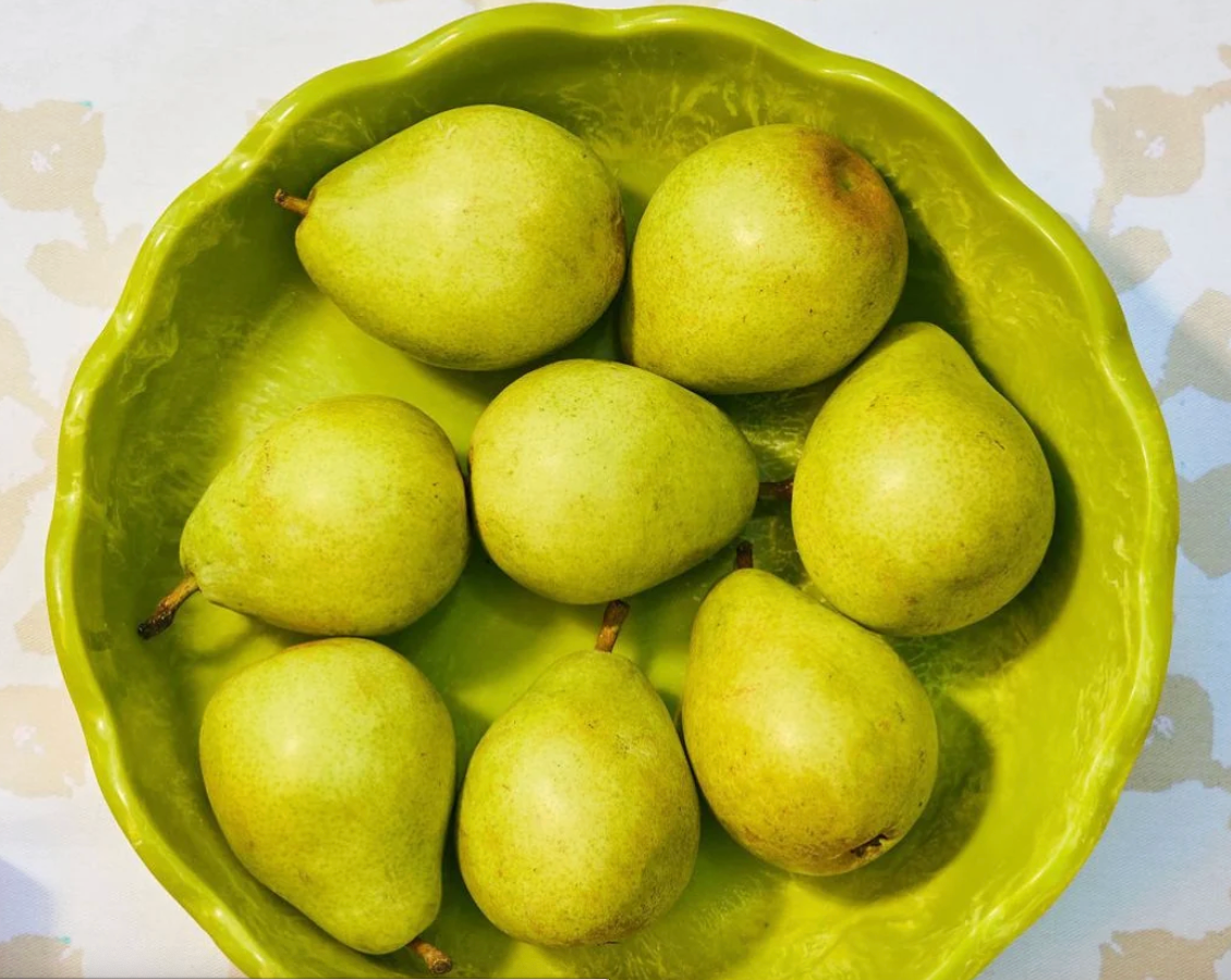 Seven green pears arranged in a scalloped lime-green resin Laura Park Designs brand bowl on a light patterned surface.