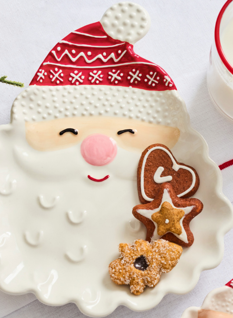 A white ceramic Christmas plate shaped like Santa's face, with a red Santa hat and white beard, styled with three gingerbread cookies on a white tablecloth.