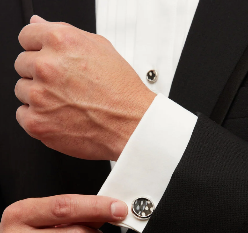 Man's hand wearing black suit and Brackish feather cuff link on a white cuff and shirt stud to match on a black suit
