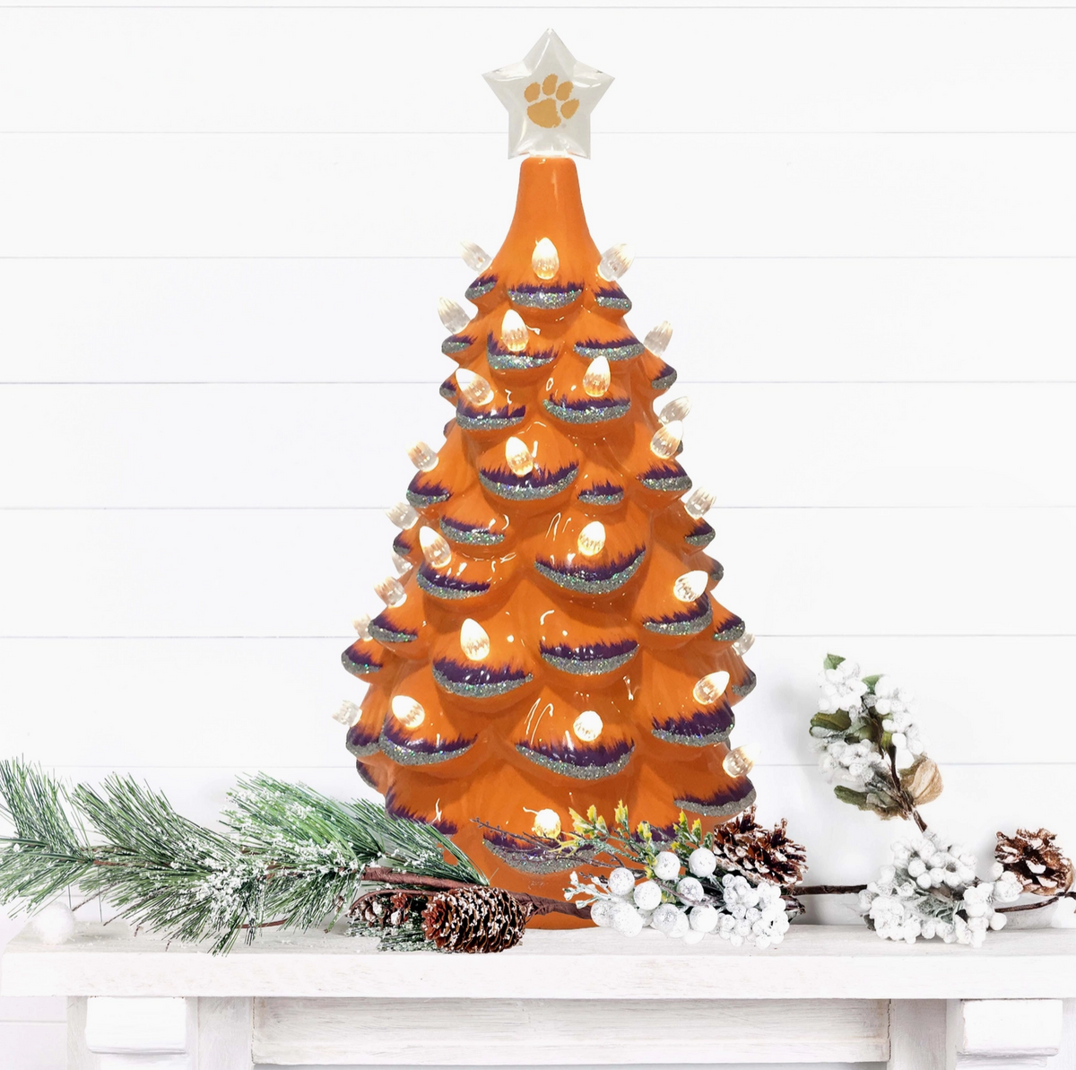 An orange ceramic Christmas tree lit and  decorated with a white star topper featuring a paw print, displayed on a white mantel with holiday greenery and pinecones against a white shiplap wall.