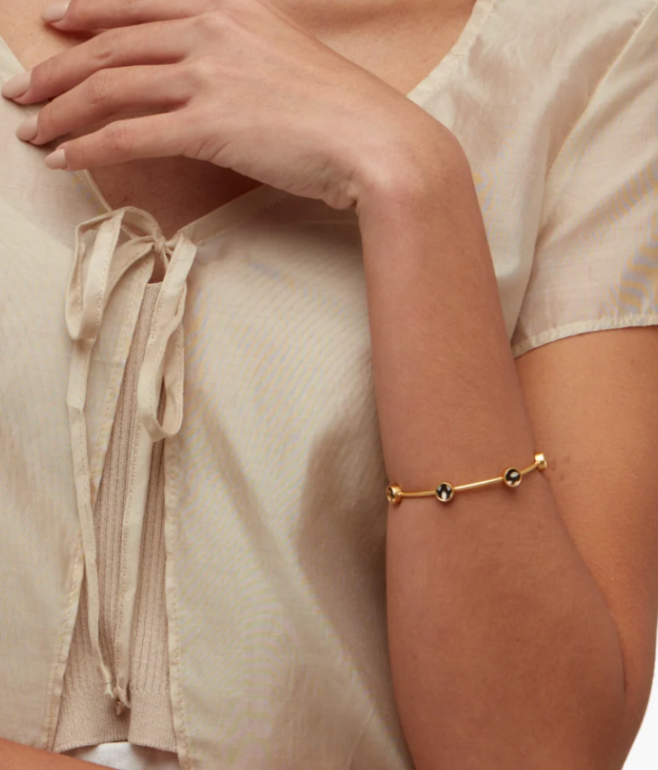 A Brackish brand gold cuff bracelet with black and white feather round inset on woman's arm with a cream blouse and bow on front