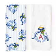 Two white kitchen towels displayed side by side on a white background. The left towel features a blue and white floral pattern with vertical light blue stripes and blue ribbon bows with green holly accents.