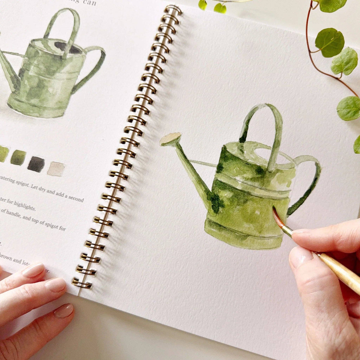 Person drawing a green watering can in a sketchbook with a real watering can and leaves on the side.