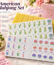 American Mahjong Set displayed on a marble surface with decorative flowers and pink accents. The set includes white tiles with colorful Chinese characters and symbols, a yellow back, and a pink storage box.