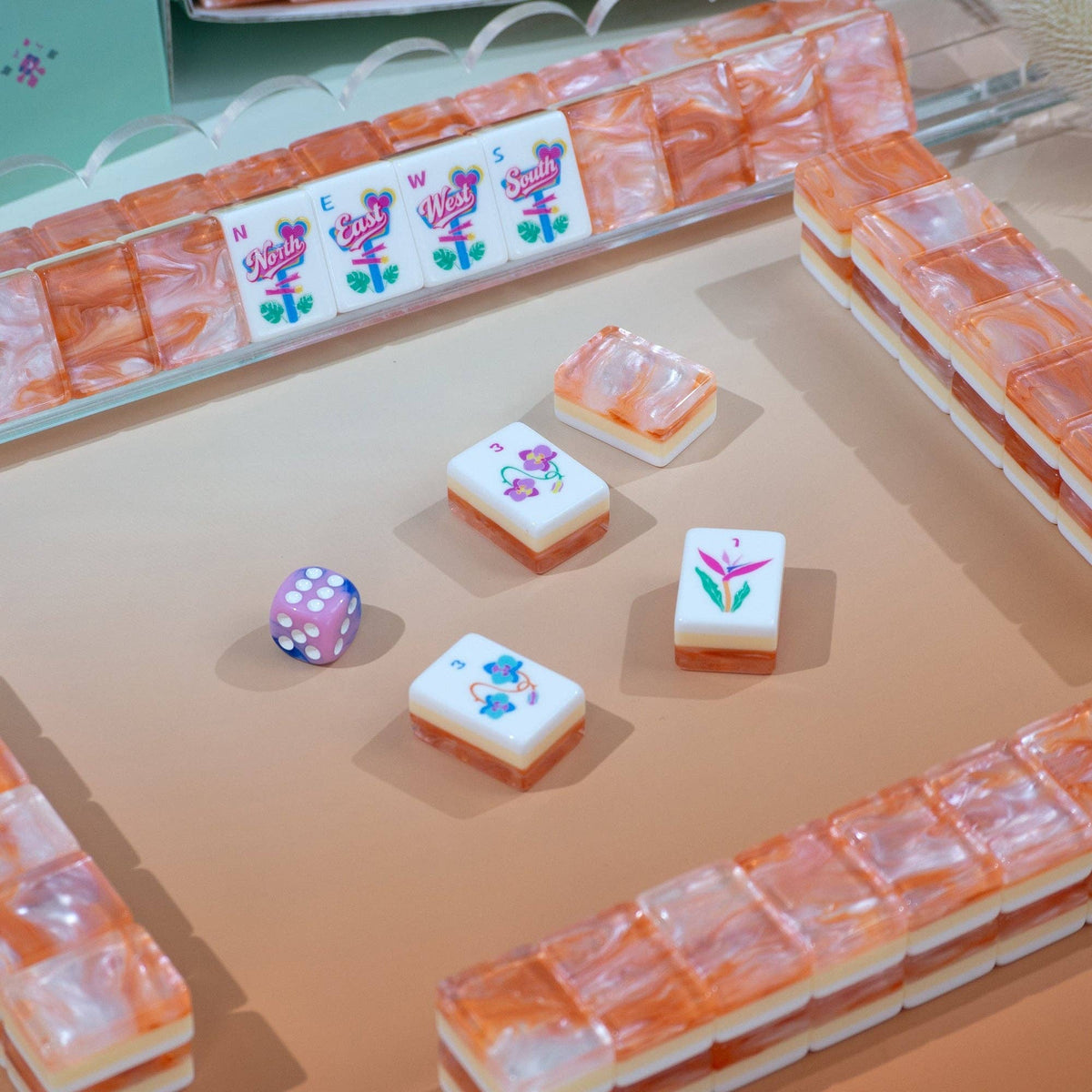 A set of mahjong tiles arranged in a game-like layout on a beige surface, with a small purple die nearby.