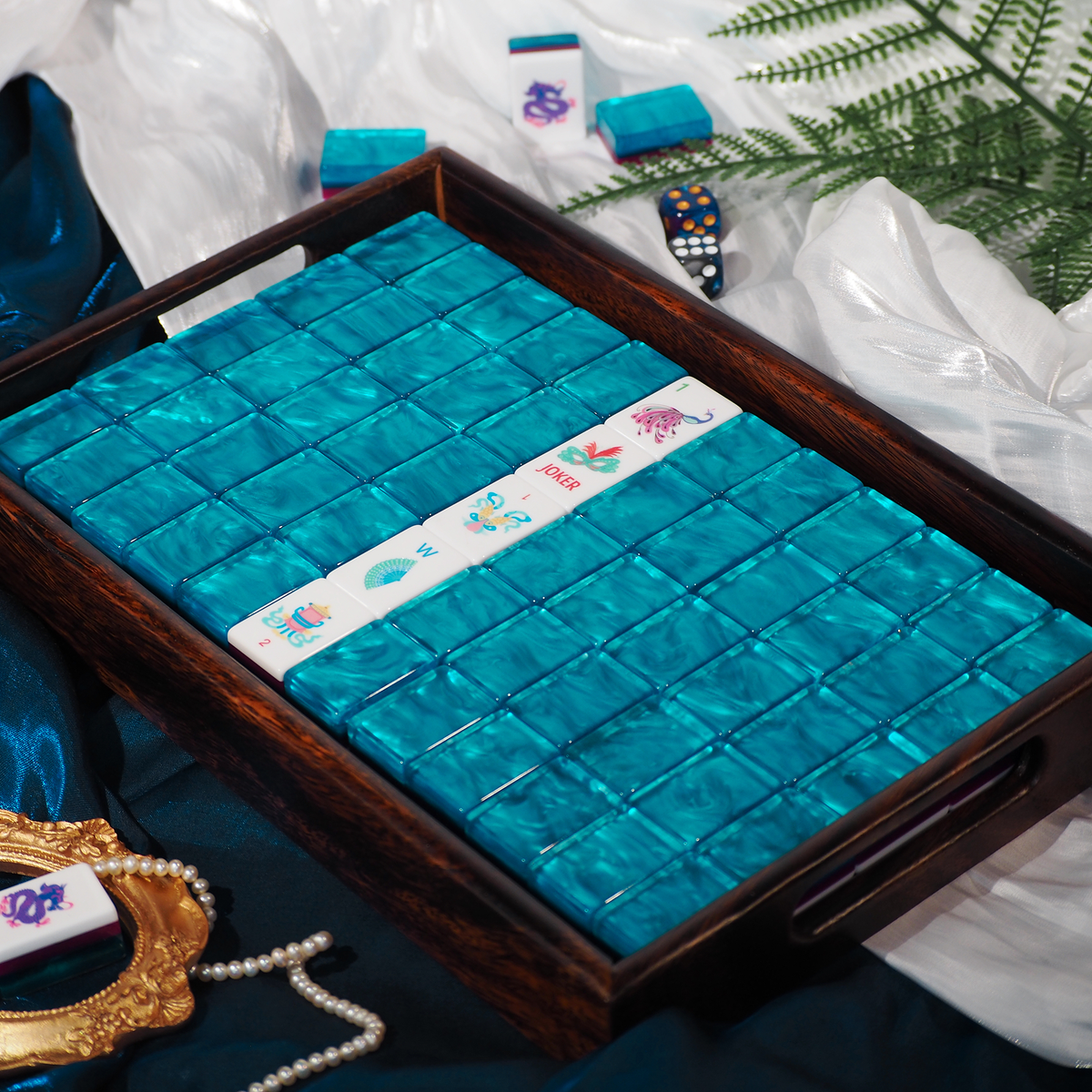 A mahjong set displayed in a dark wooden tray, featuring bright turquoise-blue backlit tiles and a white tile with colorful Chinese characters. The tray is styled on a dark blue satin fabric with white draped cloth, a pearl necklace, and green fern leaves in the background.