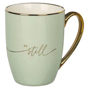 Mint green mug with gold handle and 'Be still' text on a white background
