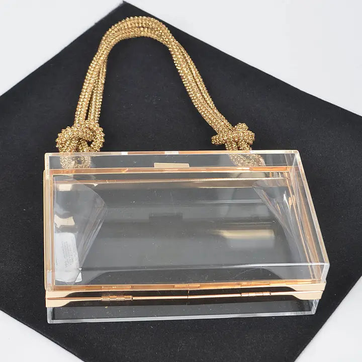 Clear acrylic box clutch with knotted gold rope top handle, open top view on black velvet showing interior capacity and gold hinge hardware