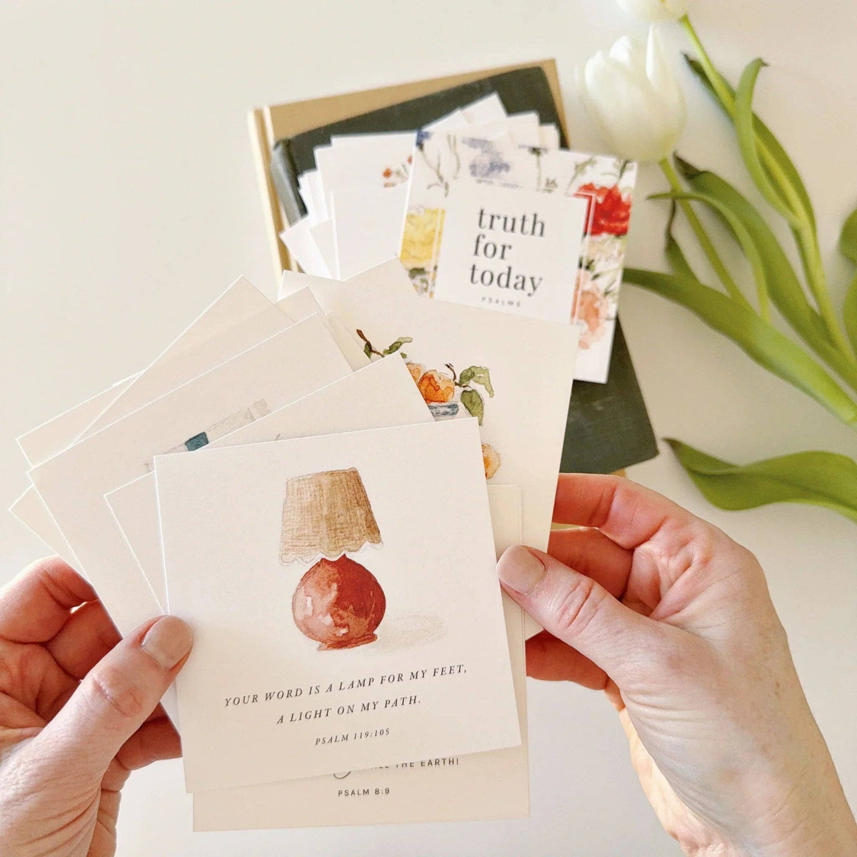 Hands holding a stack of small cards with illustrations and text, with a box of additional cards and tulips in the background.