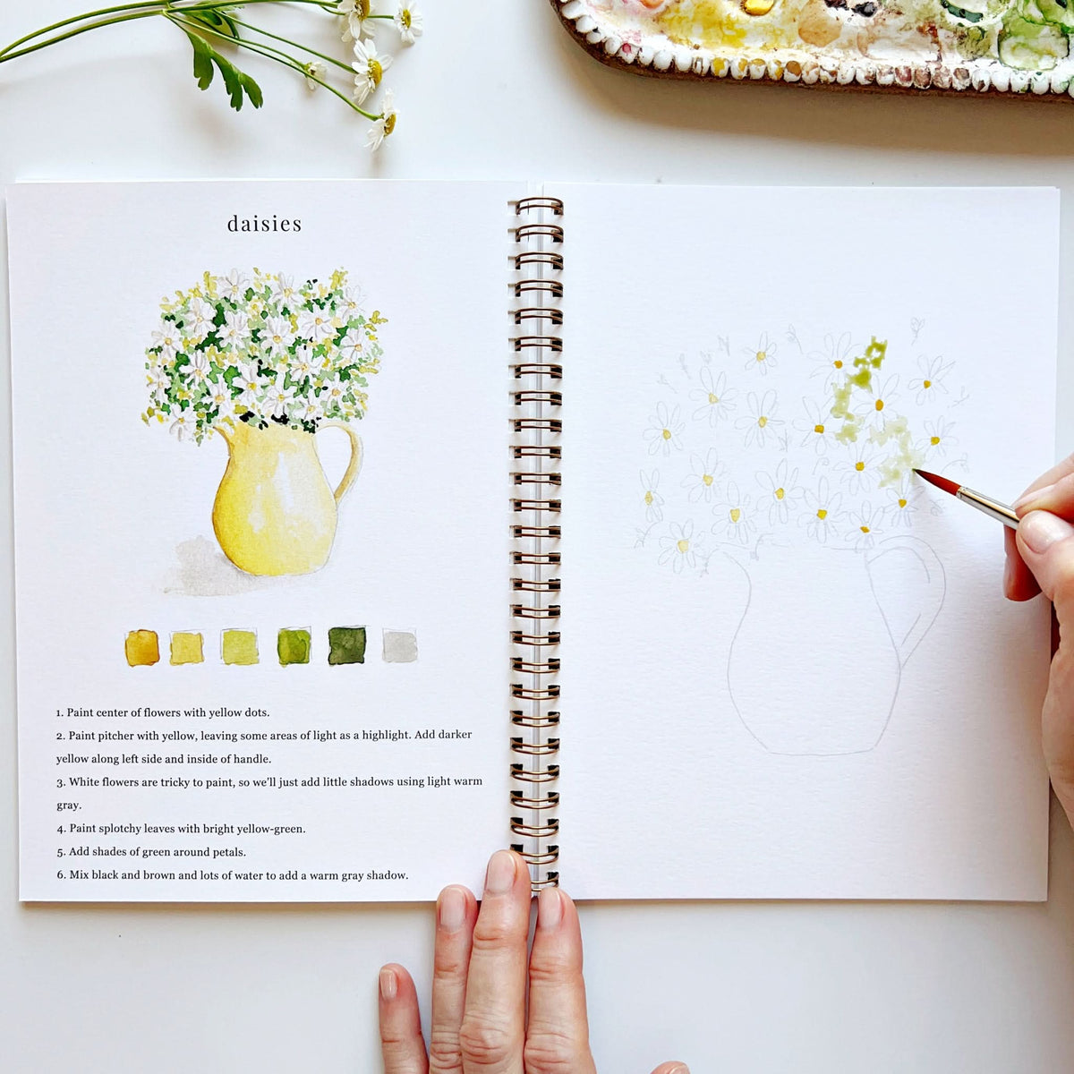 Open art book with a painting of daisies and a hand holding a paintbrush.