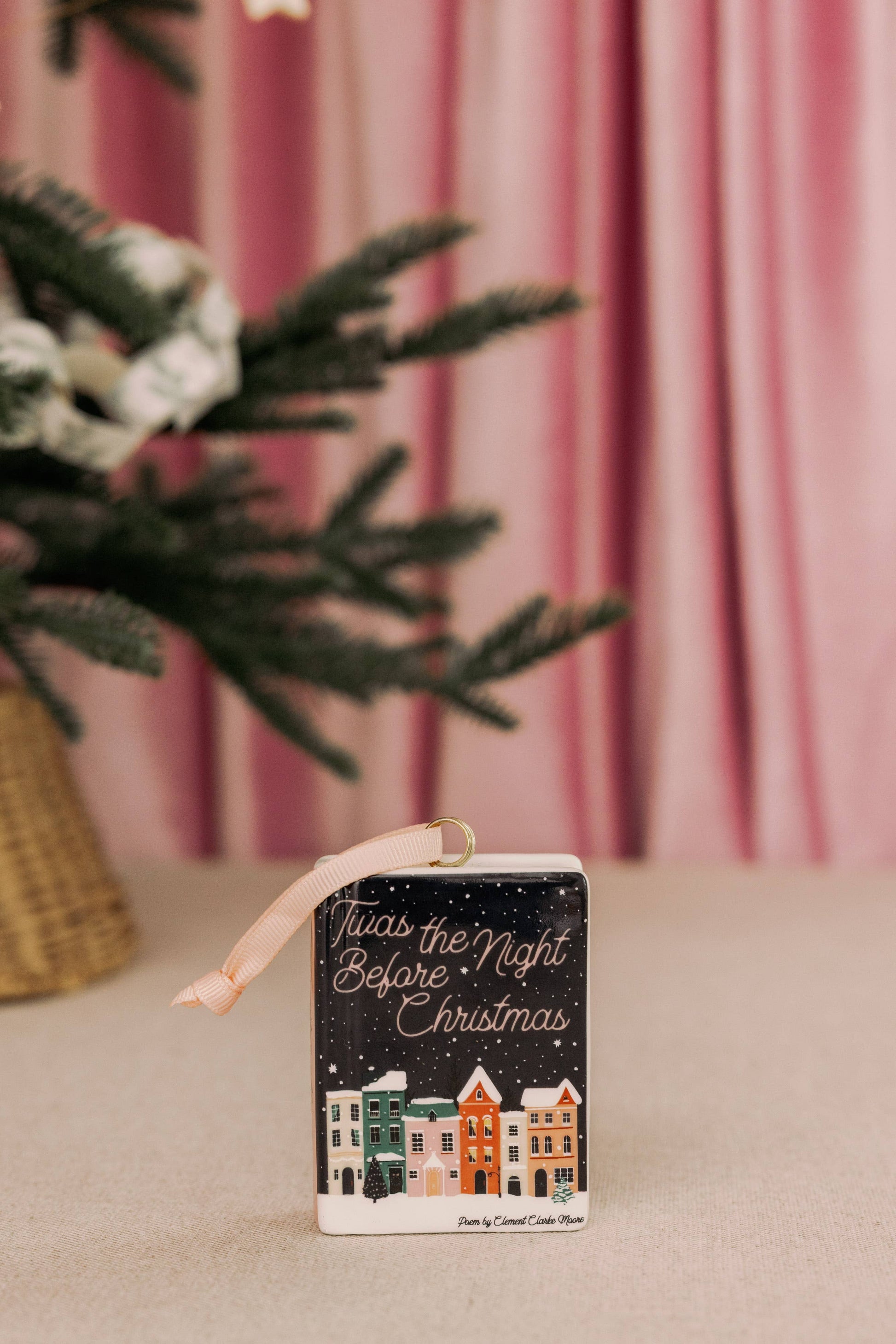 A Steel mill brand small rectangular Christmas book shaped ornament with a ribbon loop, featuring the phrase 'It was the Night Before Christmas' above a row of colorful houses under a dark starry sky. The ornament is styled on a beige surface with a pink curtain background and blurred green pine branches on the left.