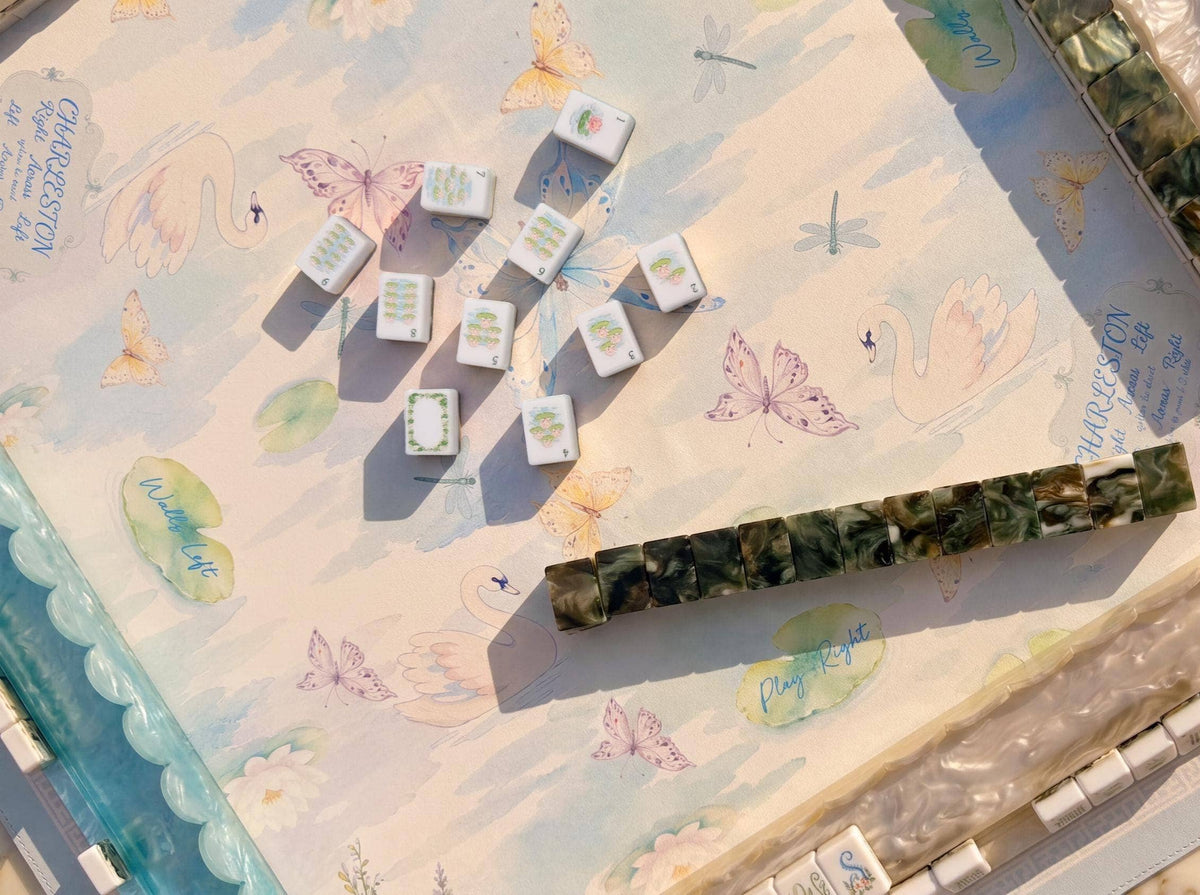 A close-up overhead view of a mahjong game setup showing several white tiles with gold characters and a long dark green marbled tile rack on a decorative pastel game board with butterflies and swans.