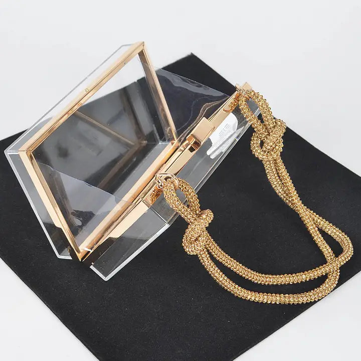 Clear acrylic box clutch open lid view showing gold frame hardware, interior mirror and knotted gold chain strap on black velvet background