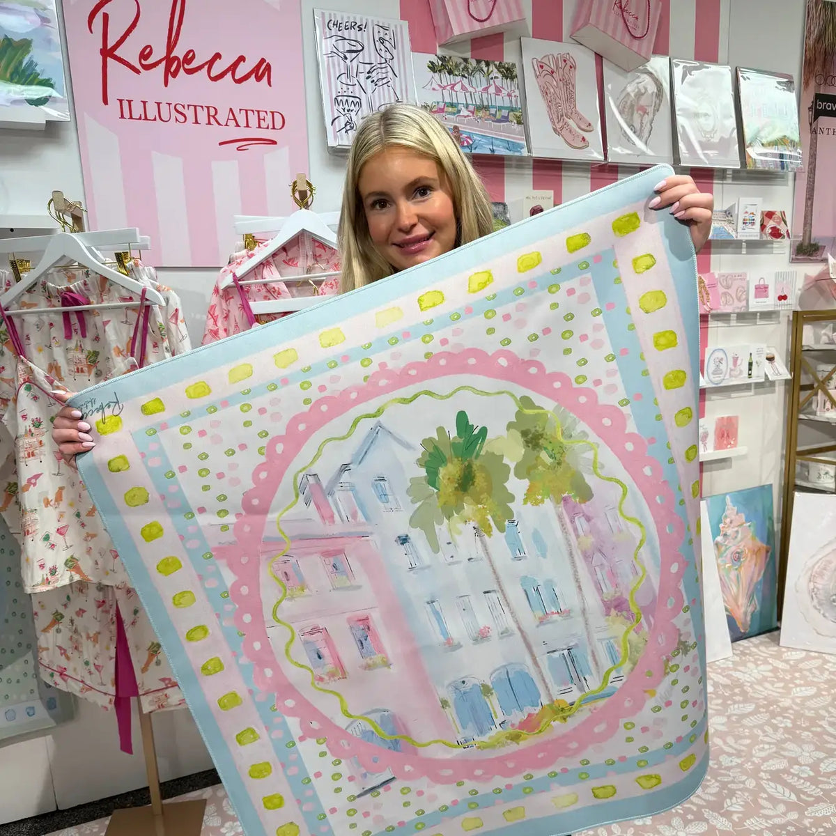 A woman holds up a large square Mahjong Mat featuring a pastel watercolor illustration of Charleston, SC Rainbow Row homes and palm trees, surrounded by pink and light blue borders with yellow accents. The background shows a boutique interior with clothing racks and a pink sign reading 'Rebecca Illustrated'