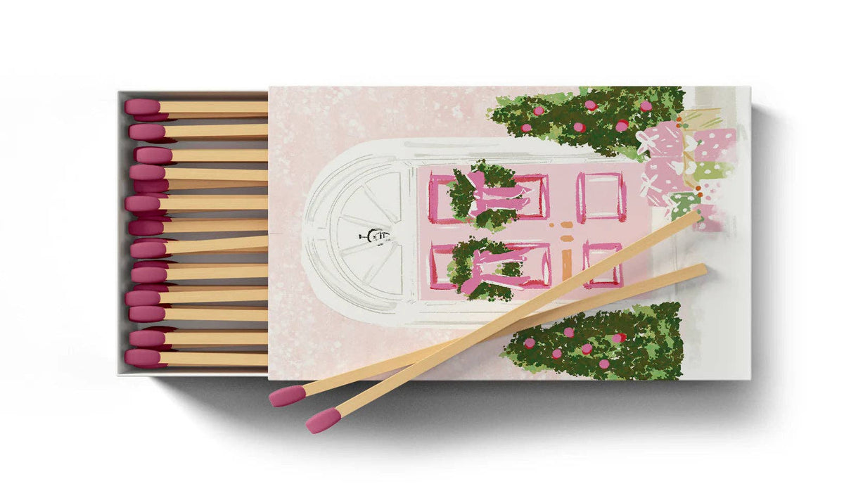 A matchbox-style decorative product shown on a white background. The box contains wooden matches with dark pink tips, and the lid features an illustrated pink door with greenery and pink flowers.