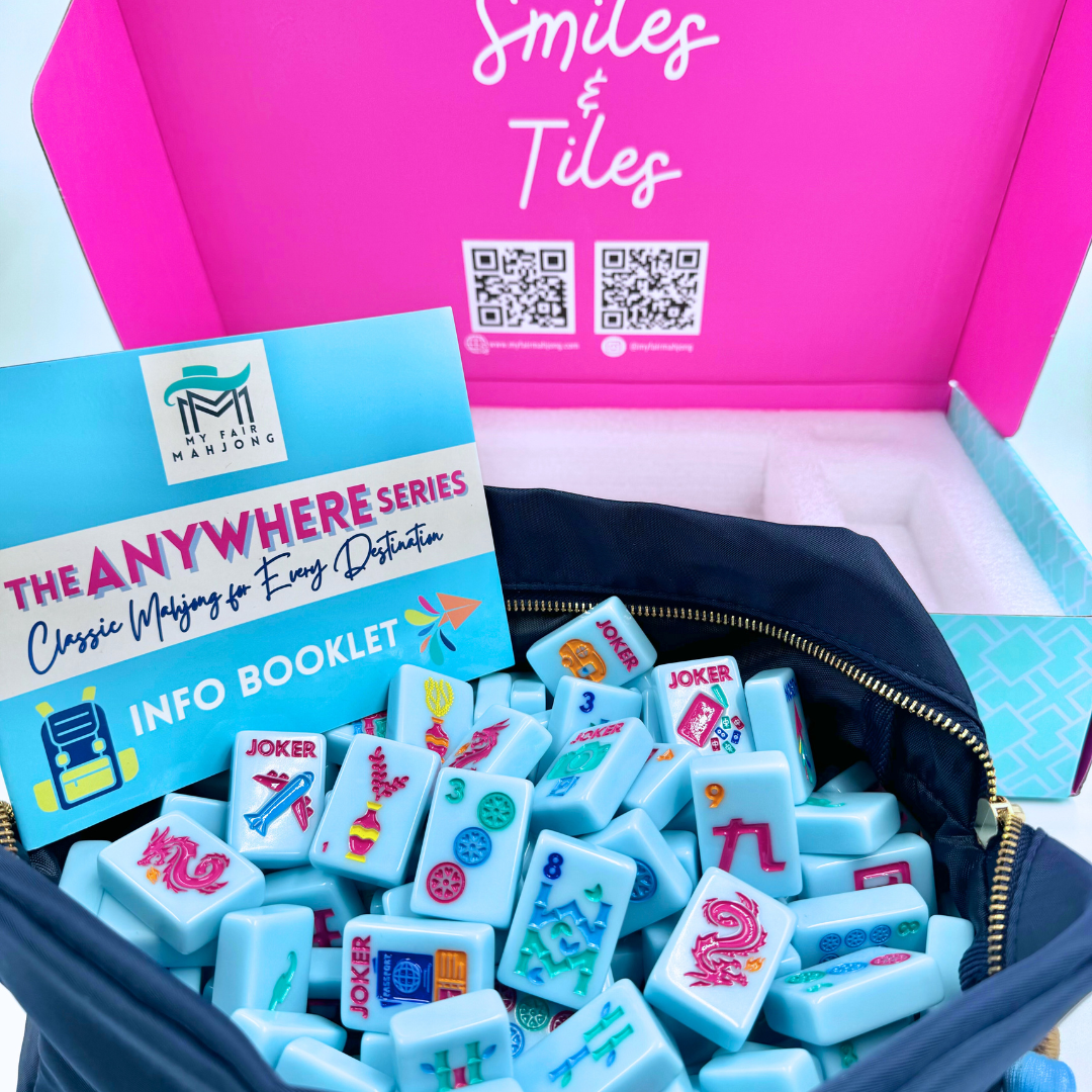A close-up of a navy zippered bag filled with light blue mahjong tiles, with a blue and white info booklet in front. A bright pink box labeled 'Smiles & Tiles' and 'The Anywhere Series Classic Mahjong For  Destination Info Booklet' sits behind the bag. Brand Is My Fair Mahjong