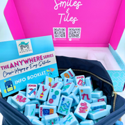 A close-up of a navy zippered bag filled with light blue mahjong tiles, with a blue and white info booklet in front. A bright pink box labeled 'Smiles & Tiles' and 'The Anywhere Series Classic Mahjong For  Destination Info Booklet' sits behind the bag. Brand Is My Fair Mahjong