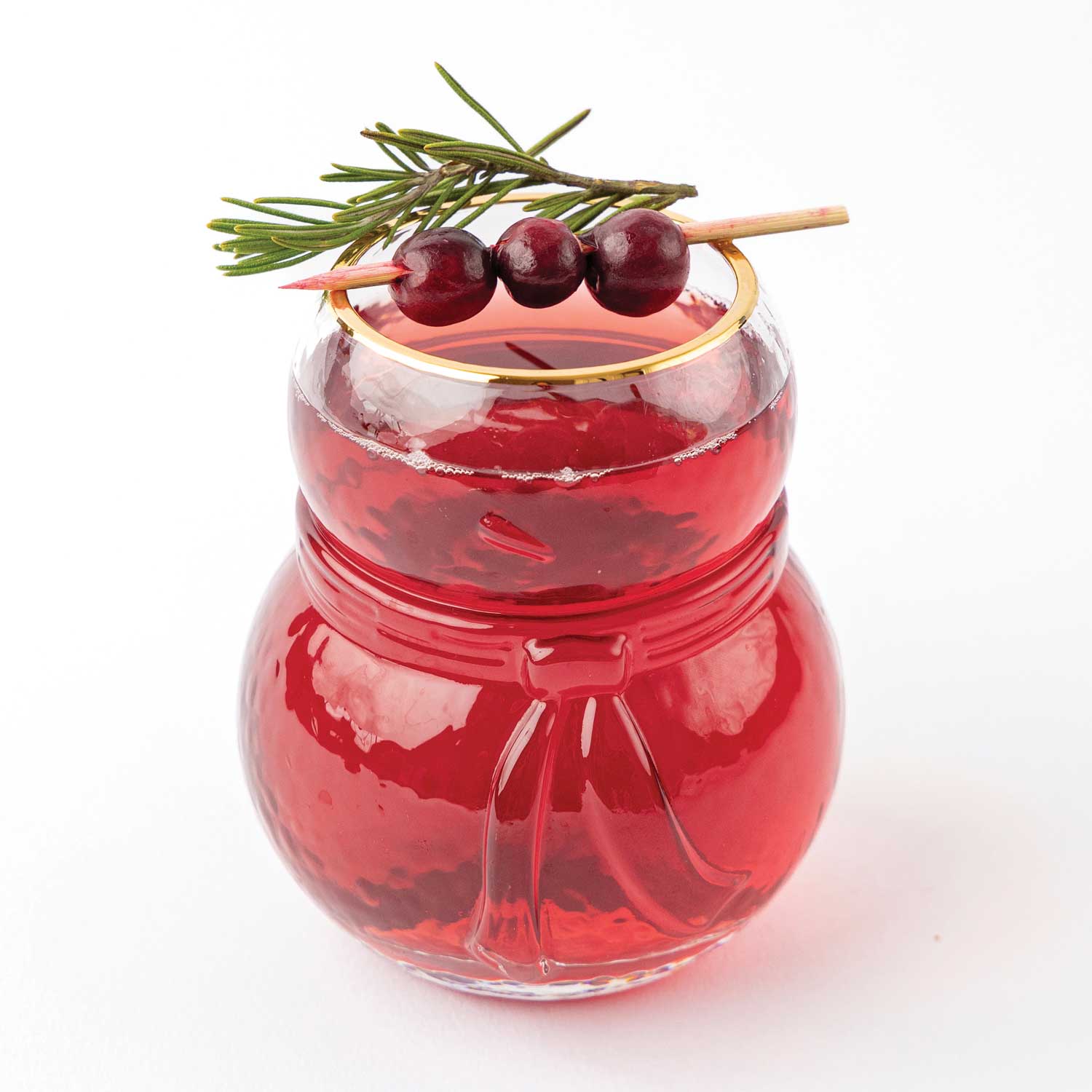 A festive 8 Oak lane brand red holiday drink in a clear snowman-shaped glass, garnished with three dark cranberries and a sprig of green pine on a wooden skewer, set against a plain white background.