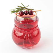 A festive 8 Oak lane brand red holiday drink in a clear snowman-shaped glass, garnished with three dark cranberries and a sprig of green pine on a wooden skewer, set against a plain white background.