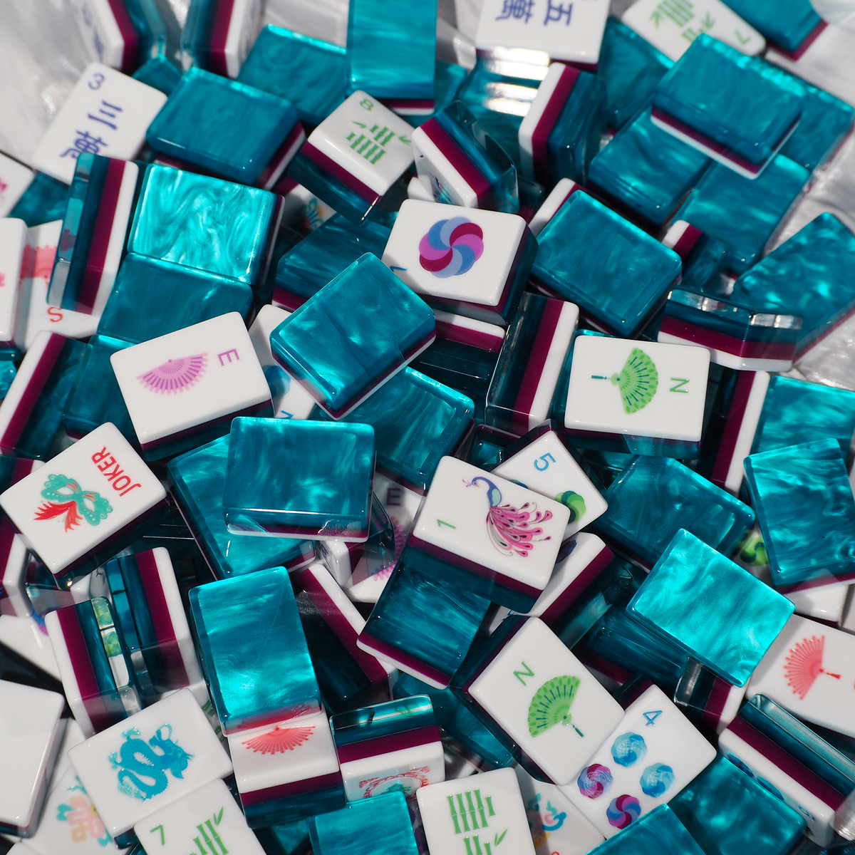 A close-up pile of Mahjong tiles with glossy turquoise blue backs and white faces showing various Chinese game symbols and illustrations.