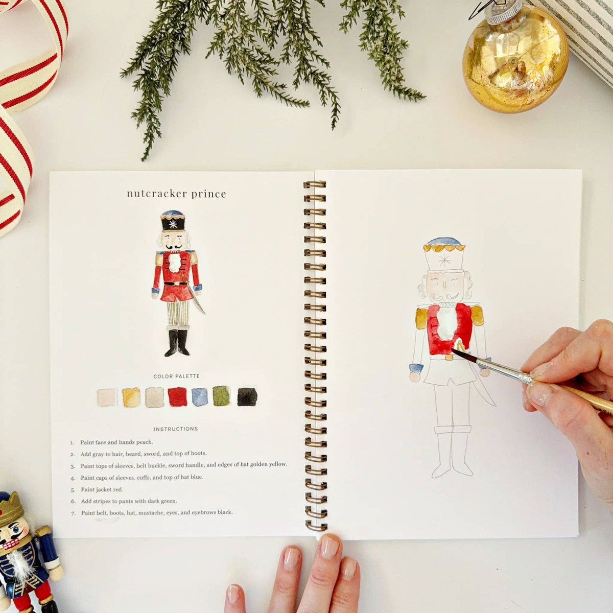 Open book with a nutcracker illustration and color swatches, hand with paintbrush paintin g the illustration and surrounded by Christmas decorations.
