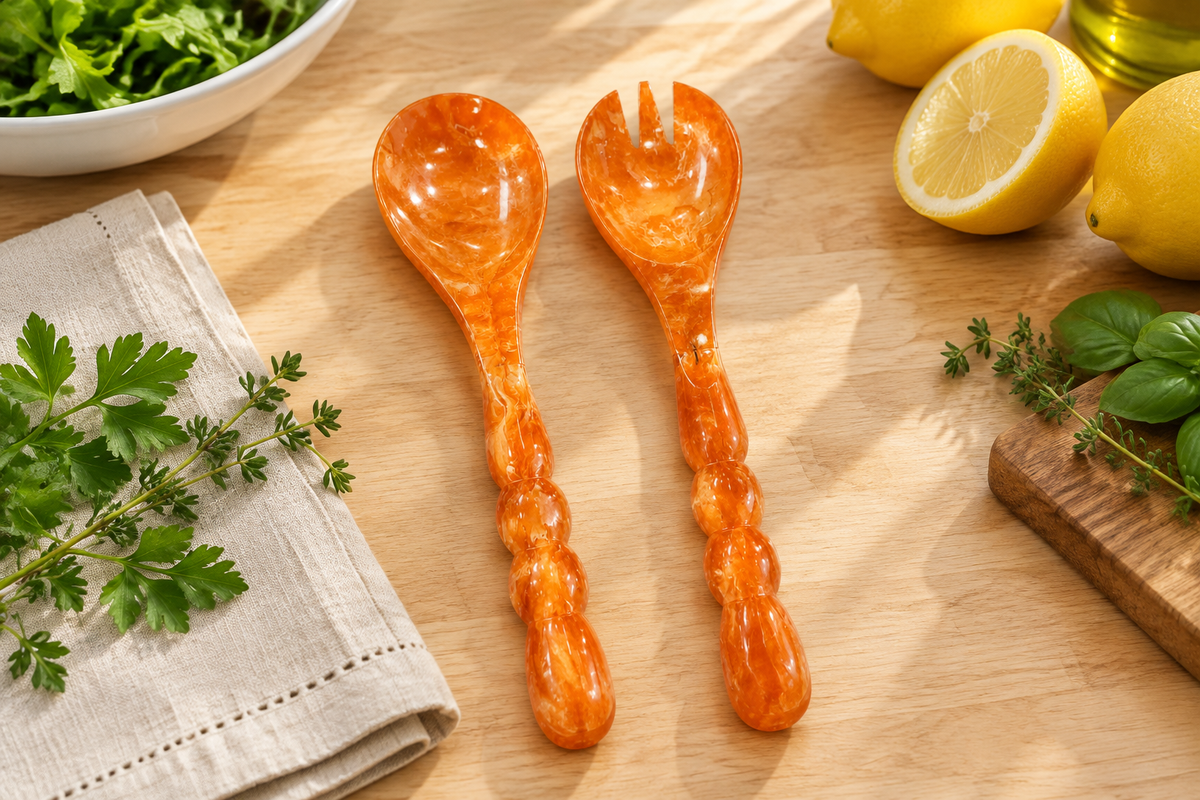 Laura Park orange marbled resin salad servers with beaded handles on a wood kitchen counter with lemons, fresh herbs and a linen napkin