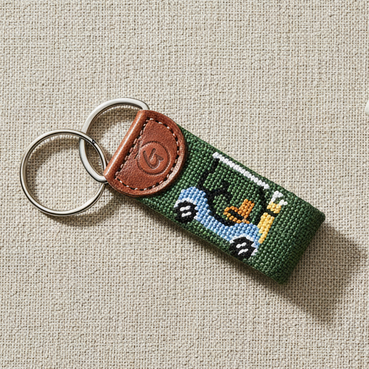 Green needlepoint golf cart keychain with brown leather fob flat lay with golf accessories
