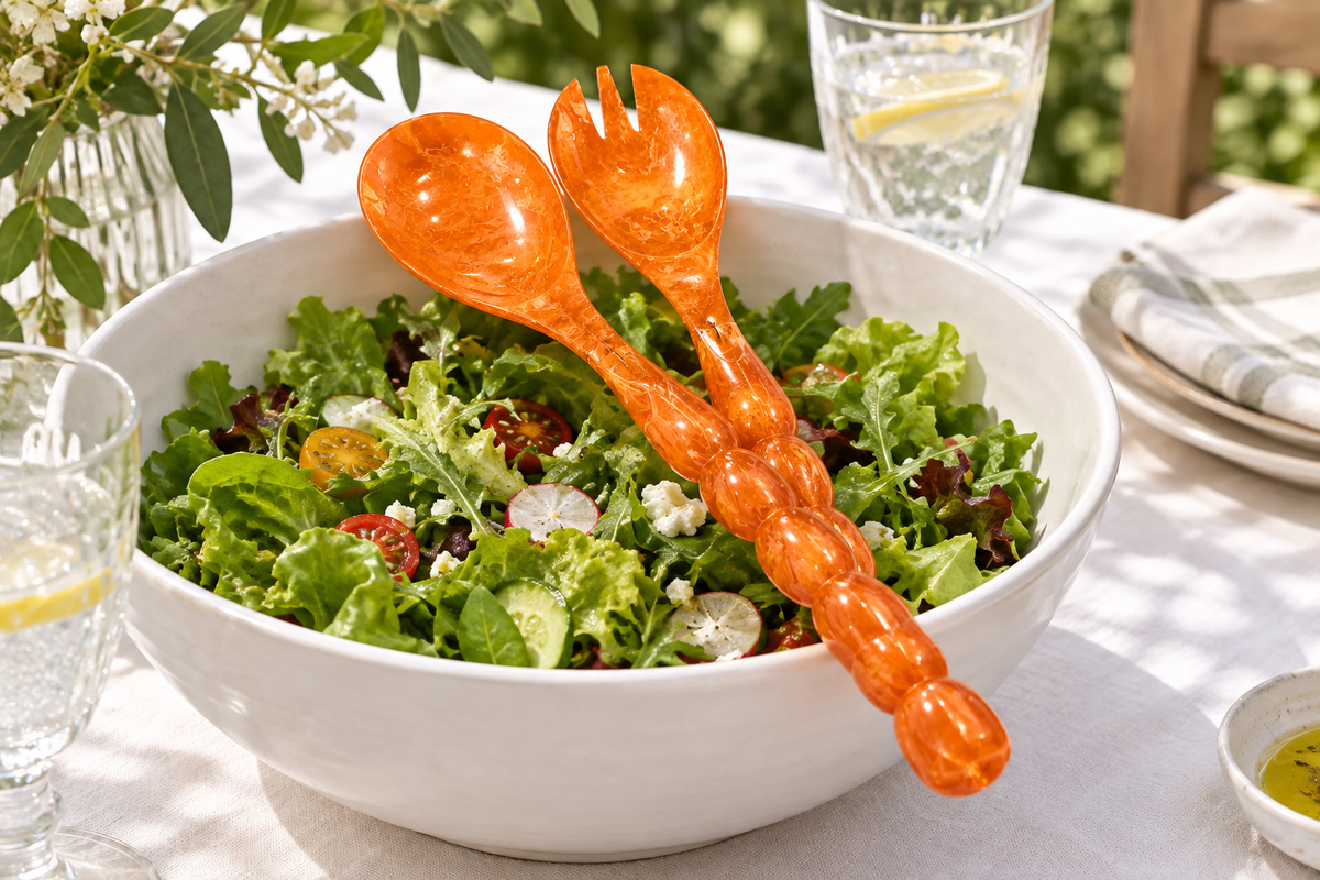Laura Park orange resin salad spoon and fork with beaded handles in a white bowl of green salad on an outdoor dinner table