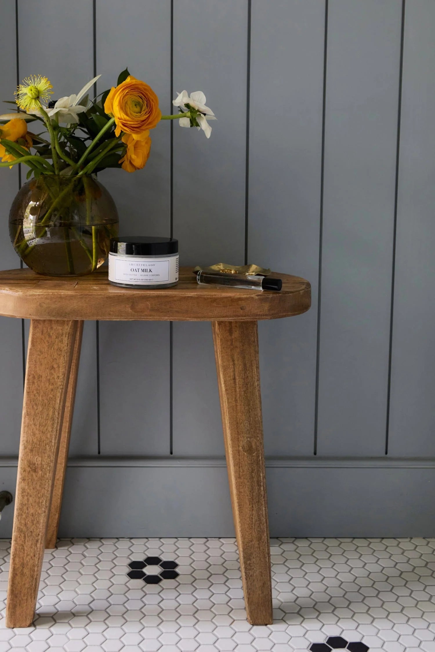 Wooden stool with a vase of flowers, candle, and small box against a gray paneled wall.