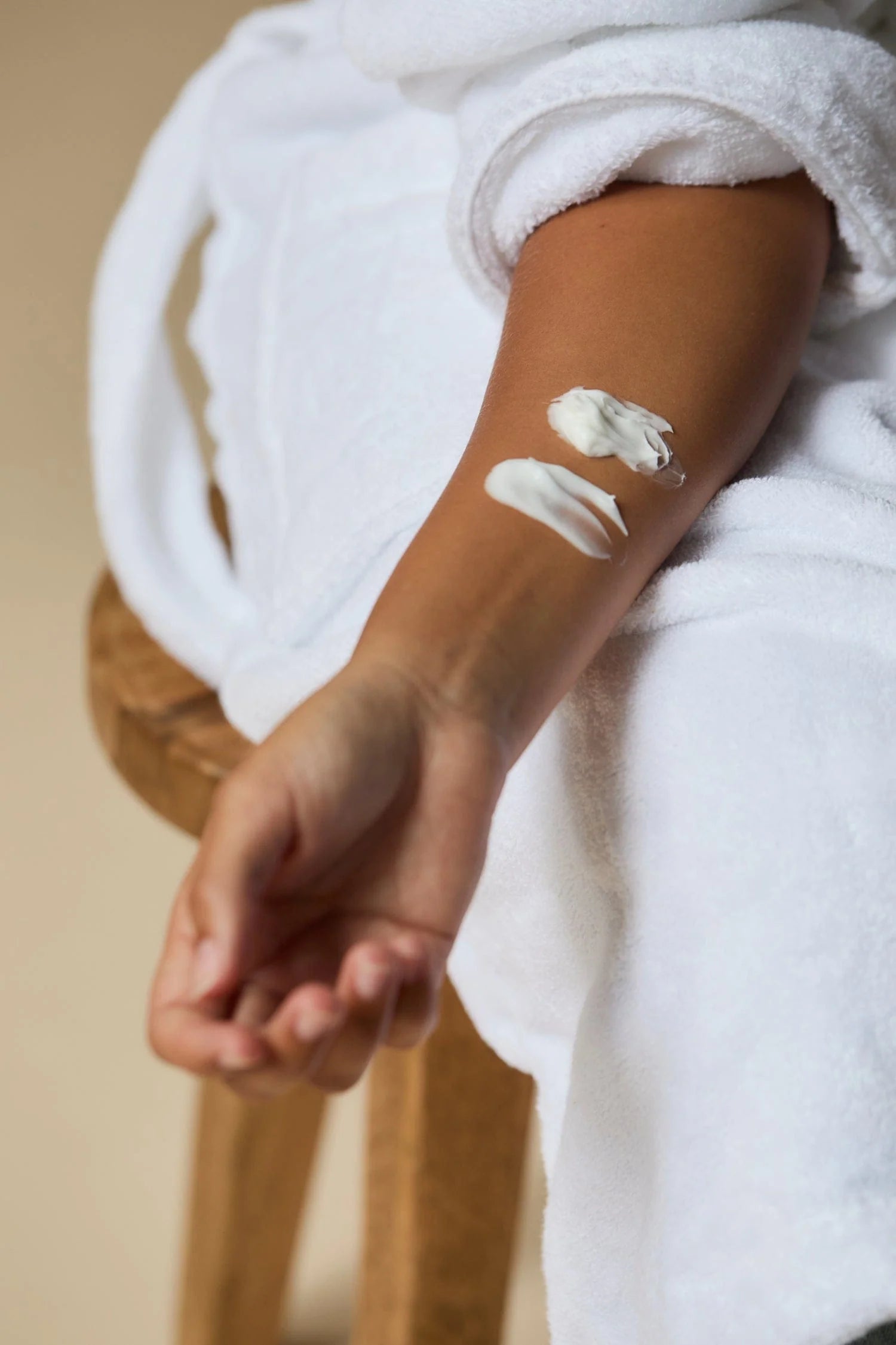 Person applying cream to their arm with a white towel draped over them, sitting on a wooden stool.